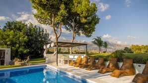 Una piscina con sillas de descanso y una pérgola, rodeada de vegetación y montañas al fondo.