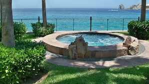 A circular hot tub with a stone border, surrounded by palm trees and greenery, overlooking a clear blue sea with a rocky outcrop in the distance. - Stunning Villa at Villa La Estancia, Directly on Medano Bay- Beautiful Views (Cabo San Lucas)