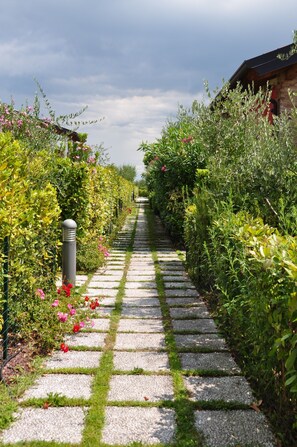 A stone pathway flanked by lush greenery and flowering plants, leading towards a building with a dark roof. - Belvedere Home a stone's throw from the center of desenzano (desenzano del garda)