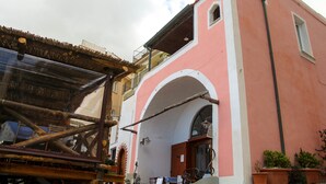 A pink building with a balcony, a wooden structure in front, and potted plants on the right.