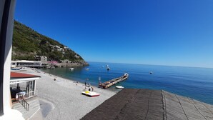 A beach with a pier extending into the sea, a rocky hillside with buildings, and a clear blue sky.