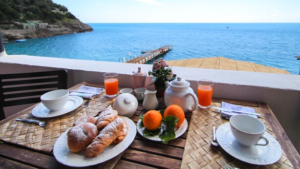 A breakfast table with pastries, fruit, and beverages, overlooking a coastal view with a pier.