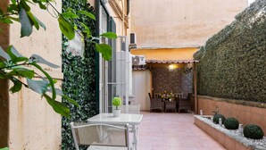 A patio area with a white table and chairs, a potted plant, and a brick wall with a tiled floor.