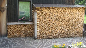 Een rustiek houten gebouw met een raam waarop roze bloemen staan, een stenen vloer en een stapel haardhout tegen de muur.