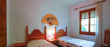 Een slaapkamer met twee houten bedden, een nachtkastje met lamp, een houten stoel, een raam met gordijnen en een decoratieve ventilator aan de muur.