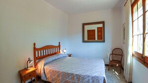 A bedroom with a wooden bed, a nightstand with a lamp, a chair, and a window with curtains. - Club Villamar - Charming villa in peaceful surroundings with private pool and fabulous sea views (Tamariu)