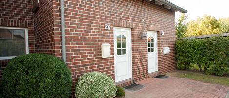 A red brick building with two white doors, numbered 24 and 25, and a small garden with trimmed bushes.