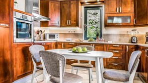 A kitchen with wooden cabinets, a white countertop, a built-in oven, a microwave, and a dining area with two chairs and a table. - Villa Xenia (Kefalonia)