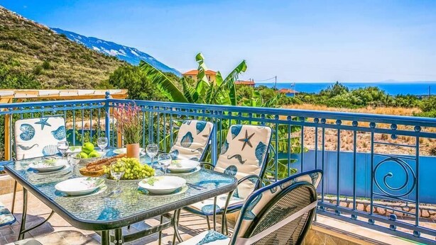 A dining area with a table set for four, outdoor chairs, and a view of the sea and mountains. - Villa Xenia (Kefalonia)