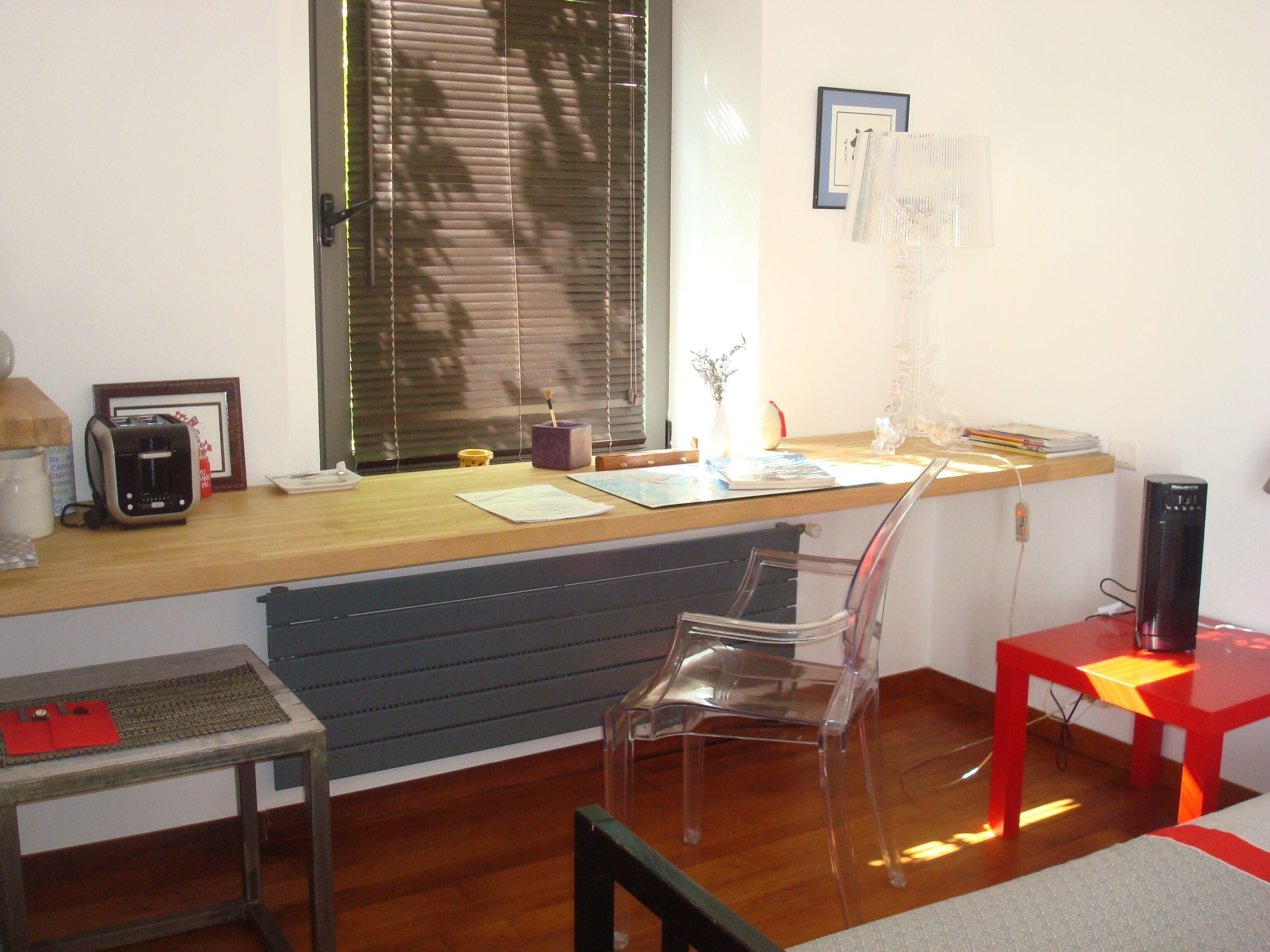 Escritório em casa com uma mesa de madeira, uma cadeira de plástico transparente, uma mesa vermelha e um radiador sob a janela.