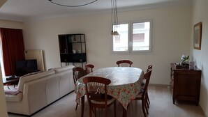 A dining area with a round table, chairs, a sofa, a TV, a bookshelf, and a window with a view of buildings. - 4-room apartment 90 m² + Terrace. Sleeps 5 Immediate proximity to beaches (Bandol)