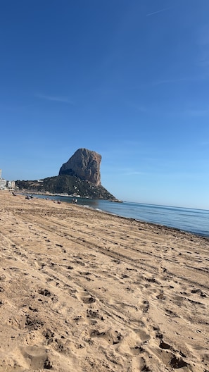 A beach with a prominent rock formation, sandy terrain, and a clear blue sky.