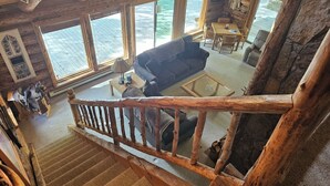 A rustic cabin interior with a wooden staircase, a living room with a sofa and coffee table, and large windows offering views of the outdoors.