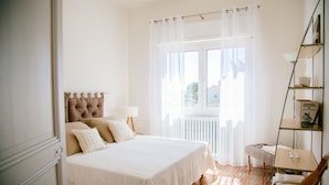 Une chambre à coucher avec un lit, une fenêtre avec des rideaux, un radiateur et une petite table avec une lampe.