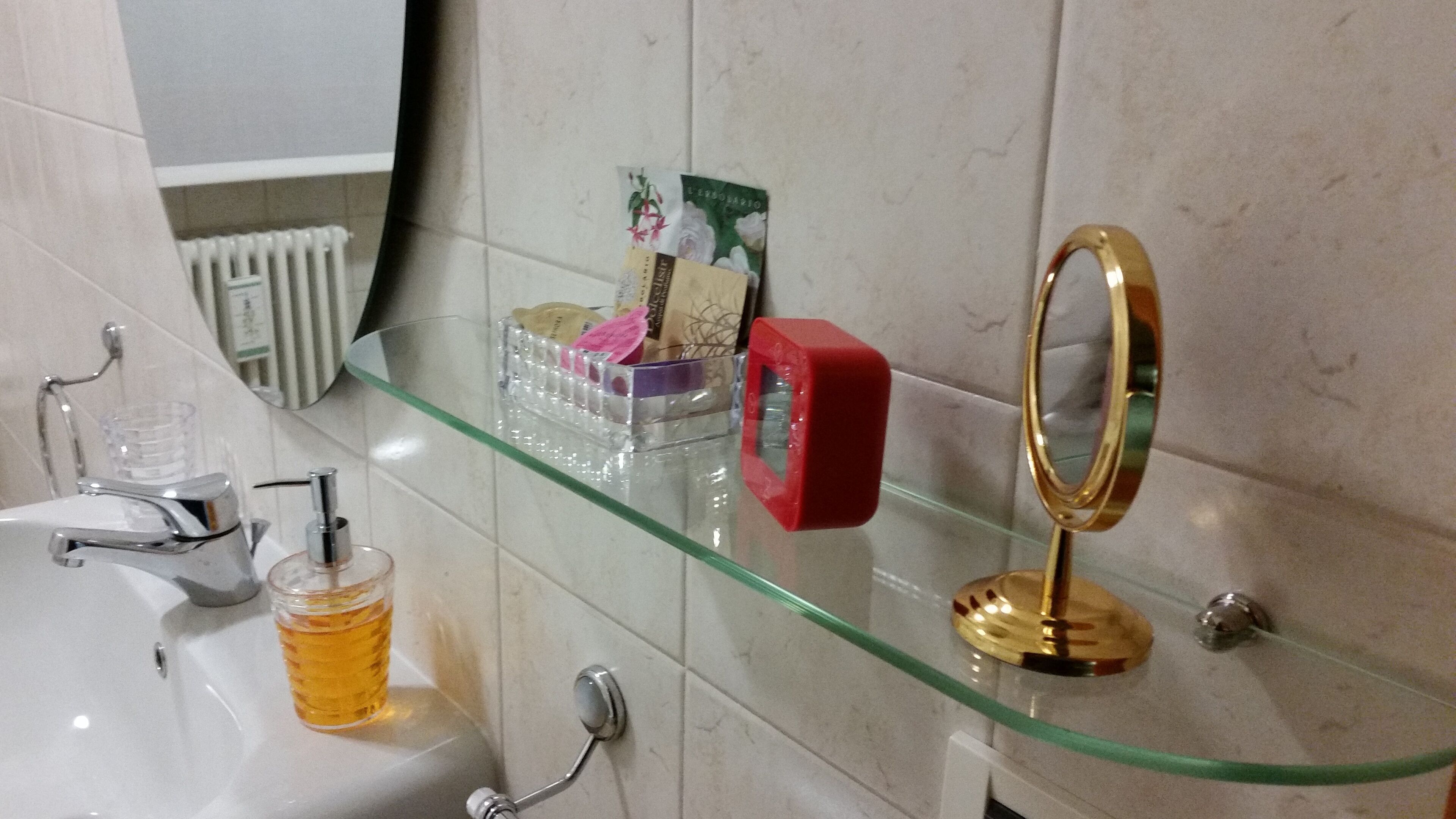Une salle de bain avec une étagère en verre où sont rangés les articles de toilette, un distributeur de savon rouge et un miroir avec un cadre doré.
