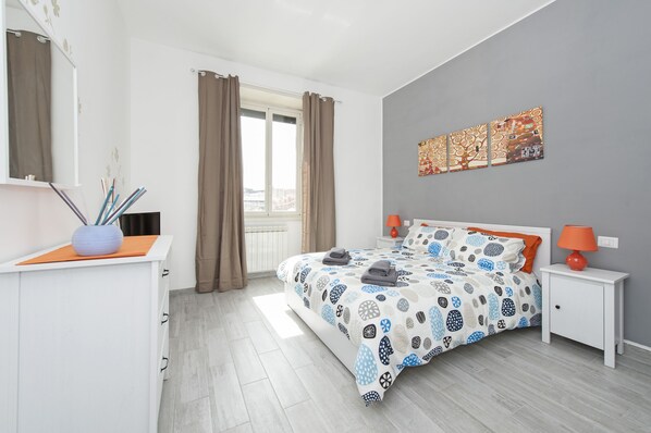 Ein Schlafzimmer mit Bett, Nachttischen, einem Schminktisch und einem Wandbild. Fenster mit Vorhängen.