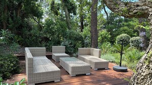 Una terraza de madera con mobiliario de mimbre, rodeada de abundante follaje y árboles.