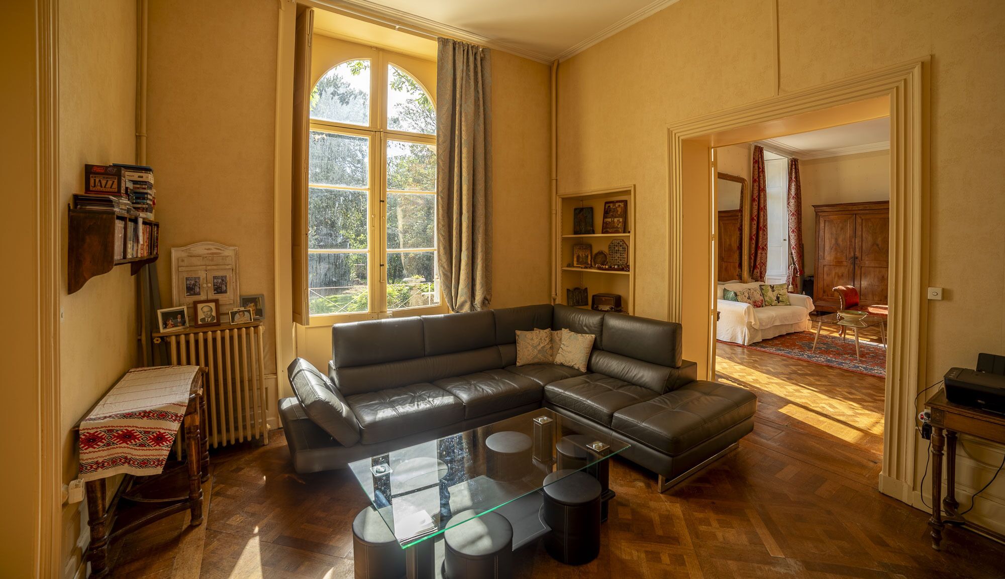 Un salon spacieux avec un canapé en cuir, une table basse en verre et une vue sur une chambre à coucher, visible à travers une porte ouverte.