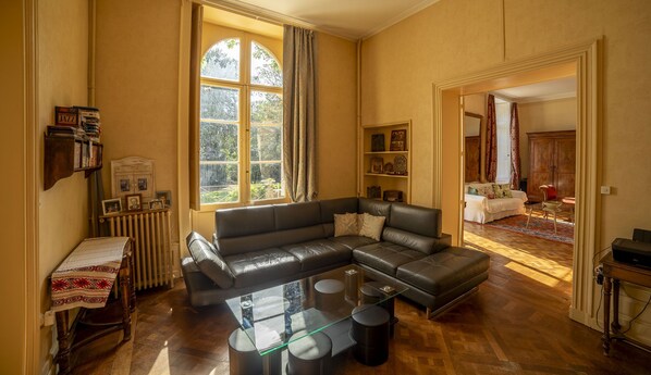 A spacious living room with a leather sofa, a glass coffee table, and a view of a bedroom through an open doorway.