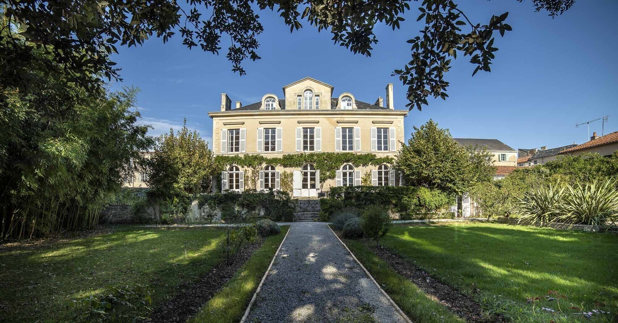 Un edificio grande e histórico con fachada simétrica, entrada central y un jardín bien cuidado con un sendero de grava que lleva a la entrada.