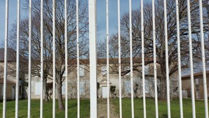 A white metal fence with vertical bars and decorative scrollwork at the bottom. Behind the fence, there is a grassy area with trees and a building with a stone facade. - House with enclosed garden adjacent cycle path leading to the beach. Internet (Dolus-D'oléron)