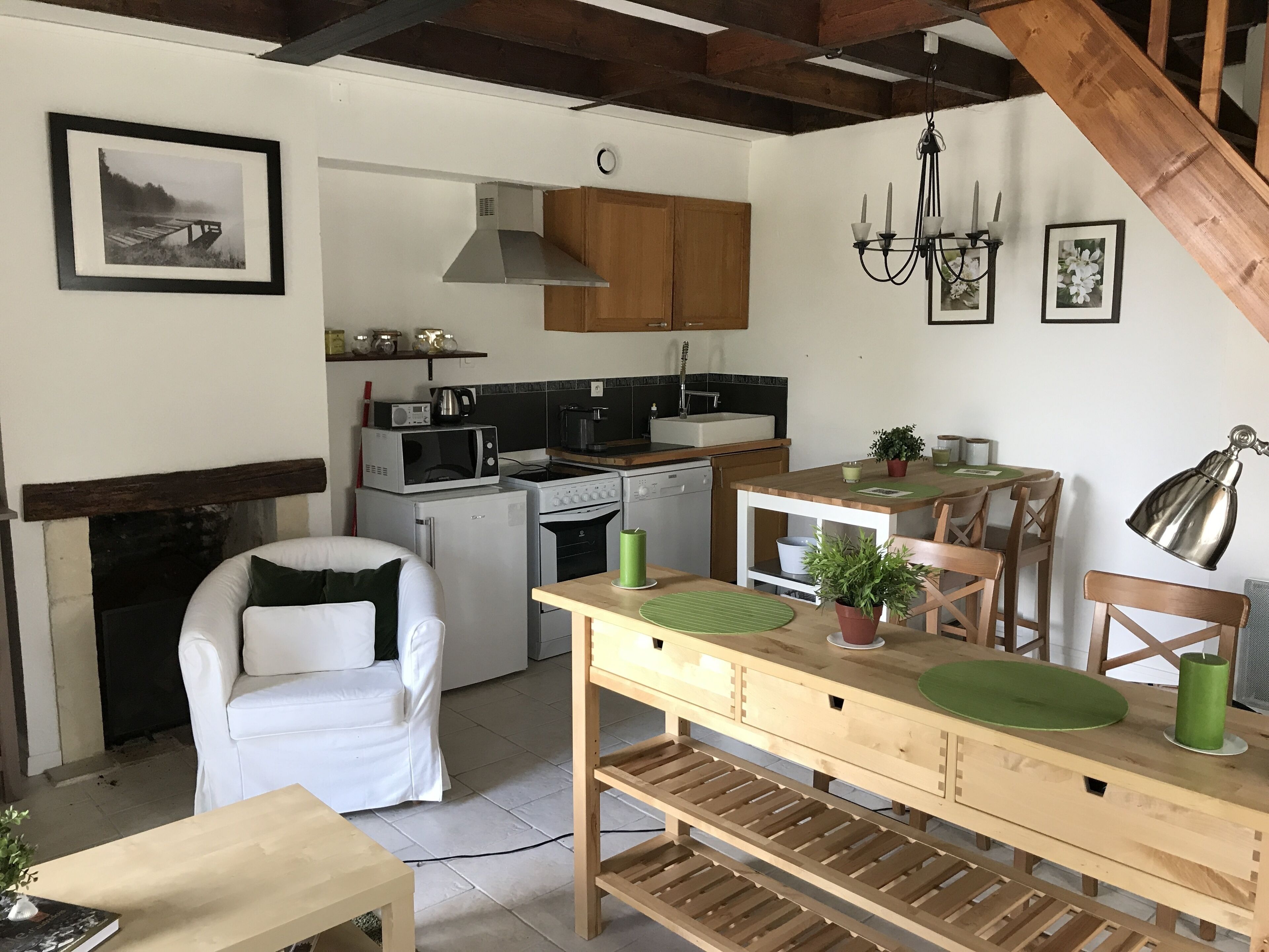 A cozy living room with a white sofa, a wooden dining table with chairs, a kitchen area with appliances, and a staircase.