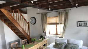 Una sala de estar acogedora con una escalera de madera, un sofá blanco con almohadones a rayas, una mesa de comedor de madera con sillas y un reloj en la pared.