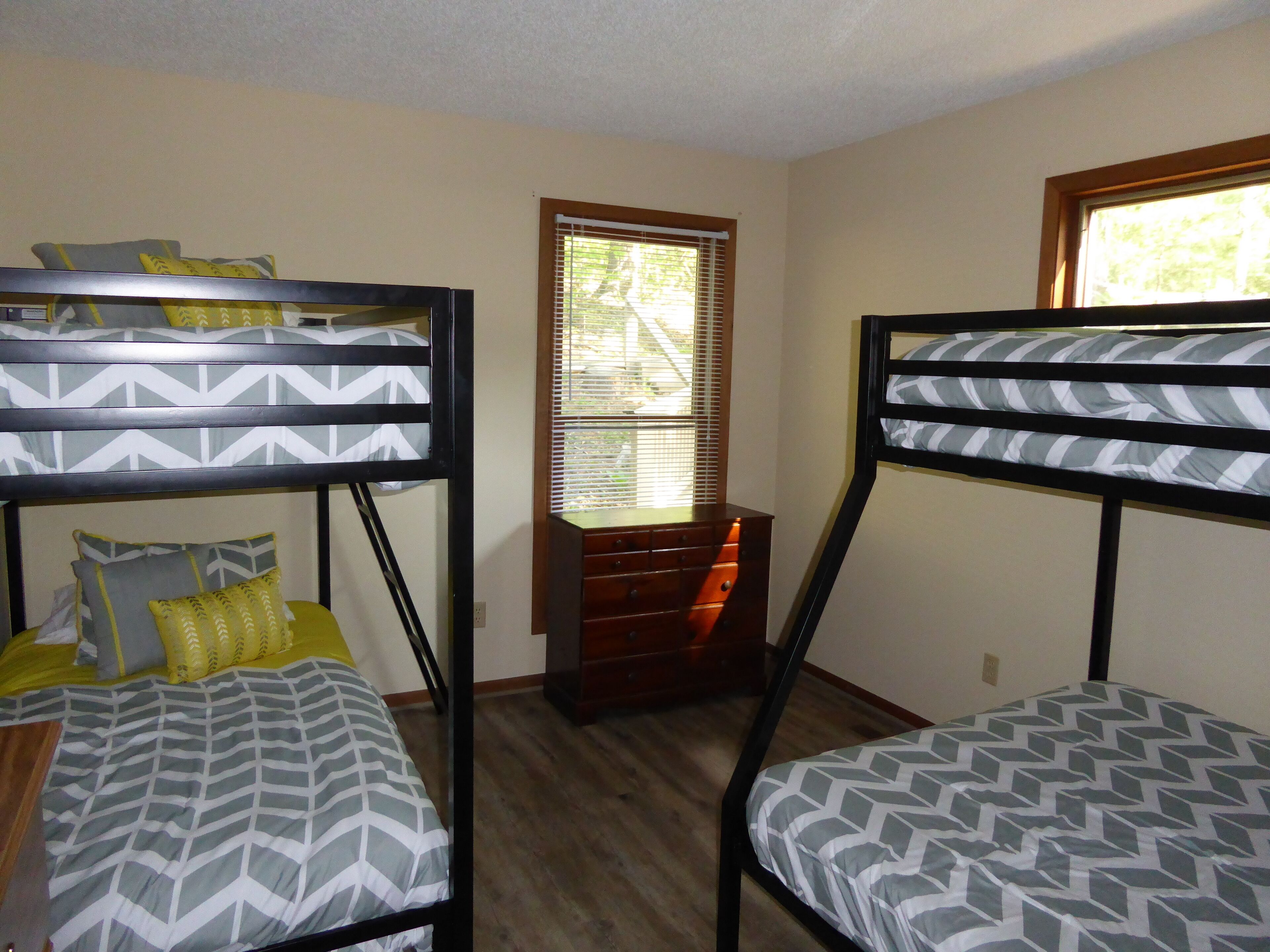Une chambre avec des lits superposés, une fenêtre avec des stores et une commode en bois.