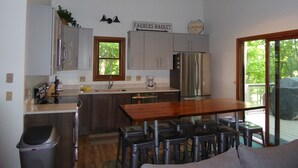 Una cocina con una mesa de madera y taburetes de bar, un refrigerador de acero inoxidable y un ventanal con vista a árboles.