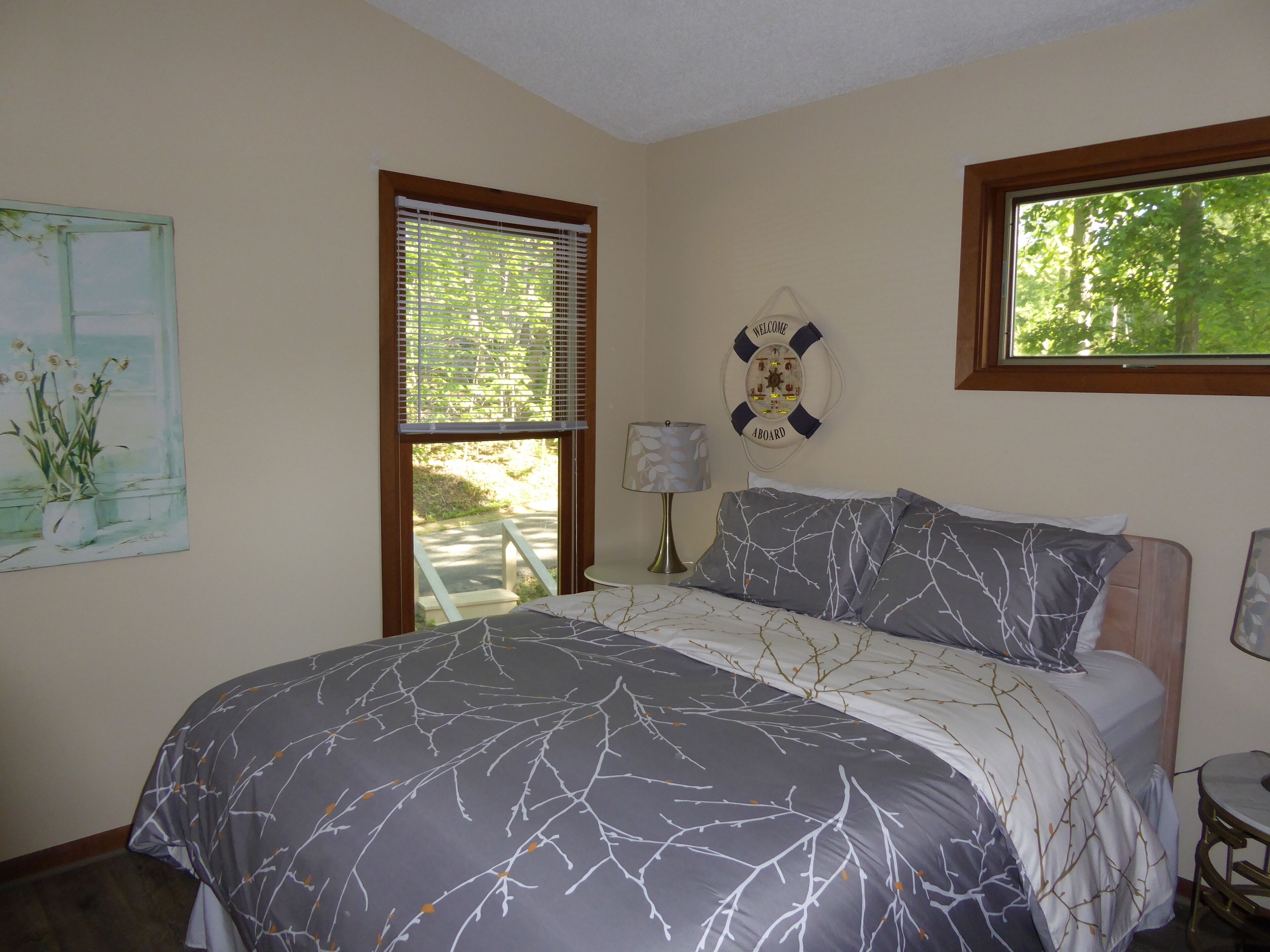 A bedroom with a bed, a window with blinds, a welcome sign, and a painting of flowers.