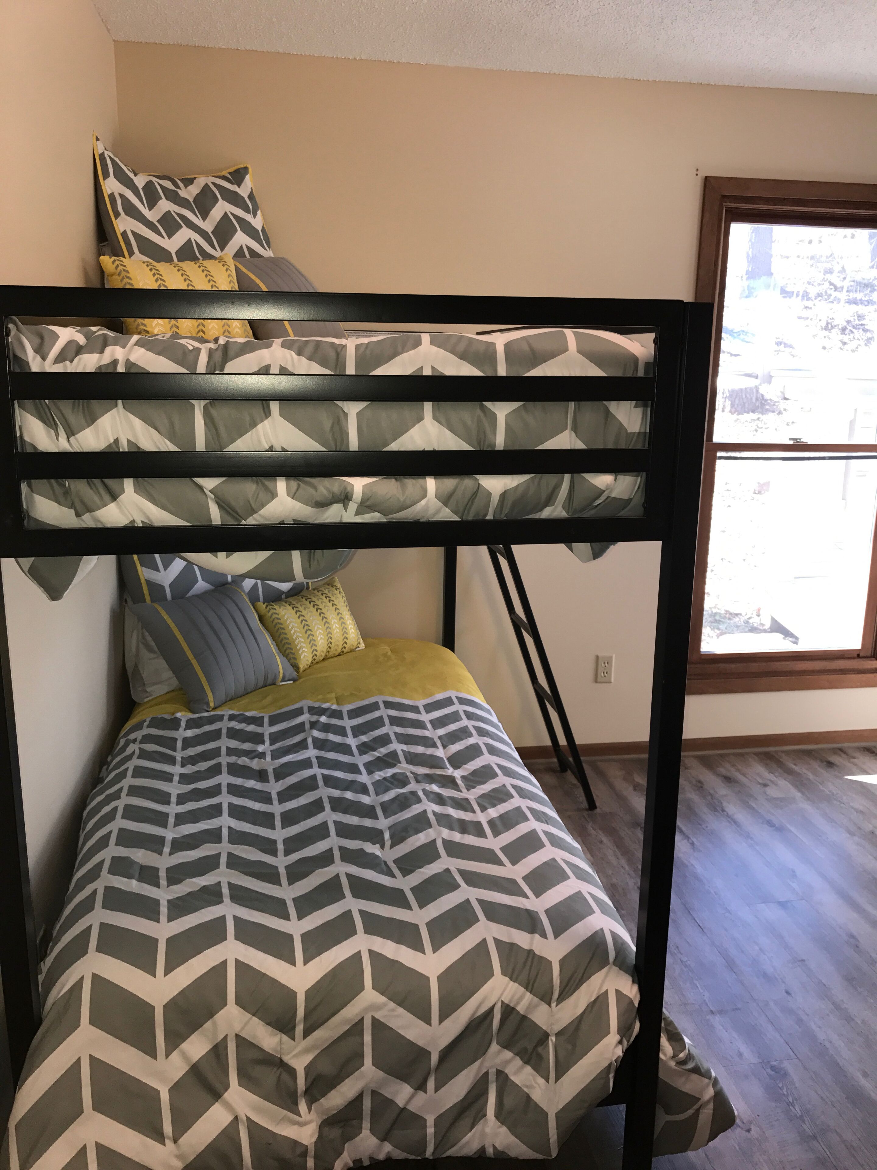 A bunk bed with a geometric patterned bedspread and pillows, a window with a view, and a wooden floor.