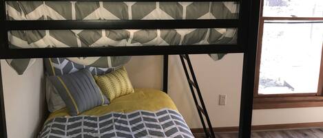 A bunk bed with a geometric patterned bedspread and pillows, a window with a view, and a wooden floor.