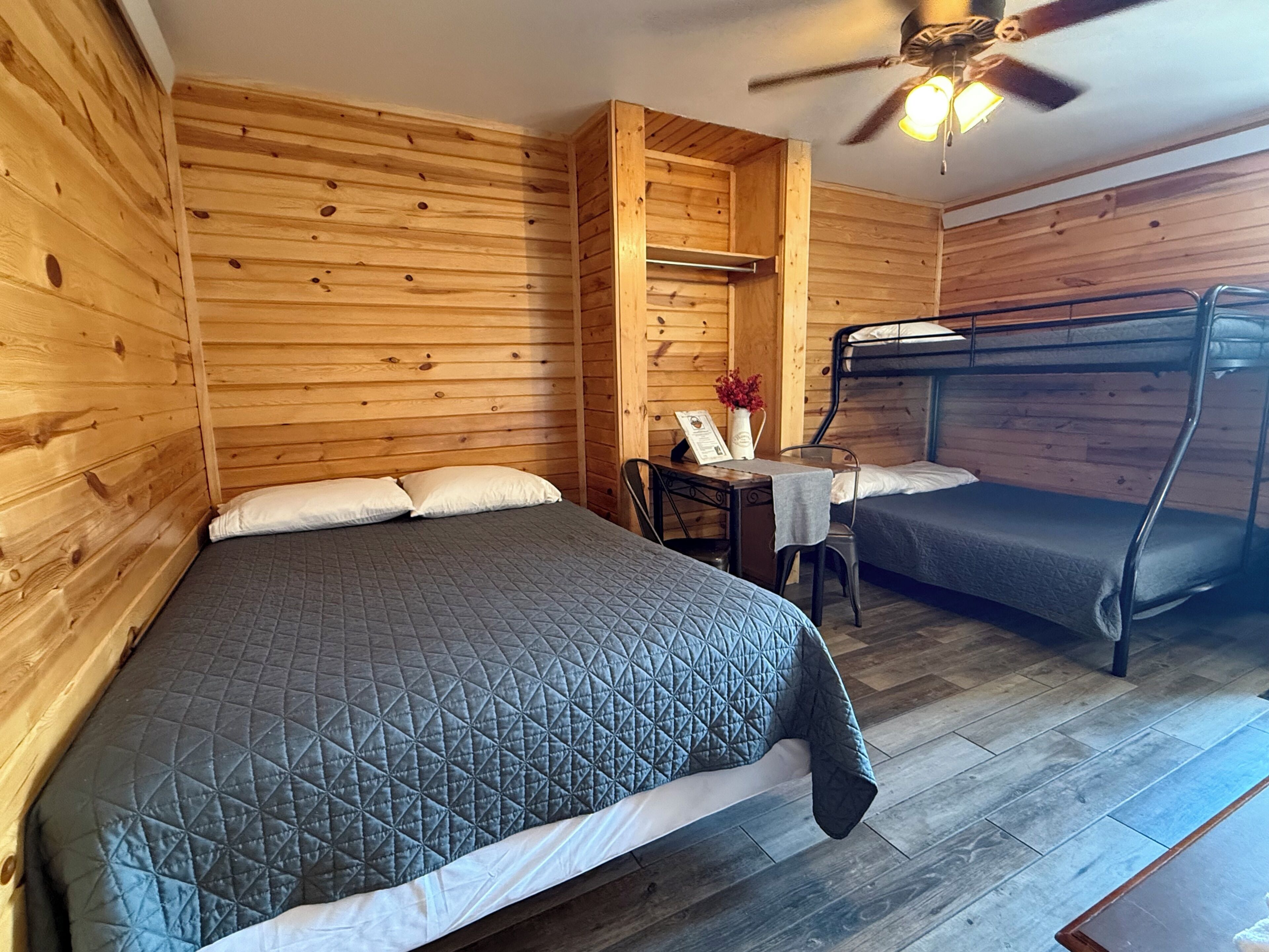 A wooden cabin room with a bunk bed, a single bed, a table with a vase, and a ceiling fan.