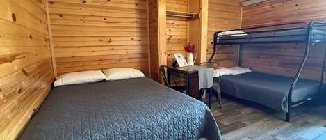 A wooden cabin room with a bunk bed, a single bed, a table with a vase, and a ceiling fan.