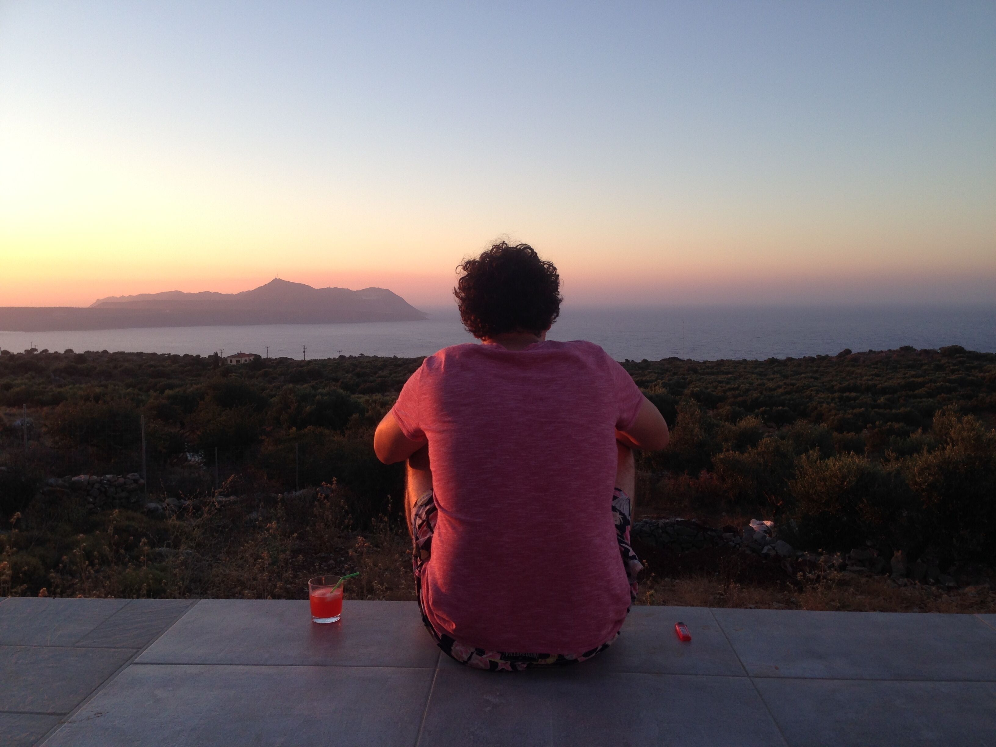 A person sitting on a tiled patio overlooking a coastal landscape with vegetation and distant mountains.