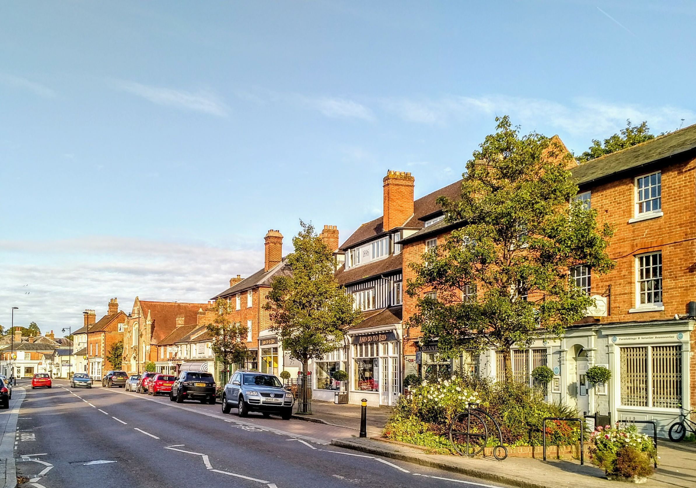A street lined with traditional brick houses, some with shop fronts, and a clear road with cars parked on the side.