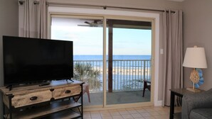 A room with a TV on a wooden cabinet, a chair, a lamp, and a balcony with a view of the beach. - Beachfront Renovated Condo | Walkable to Dining, Shopping, & Family Fun! | Pool (Gulf Shores)