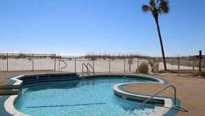 A swimming pool with a curved edge, surrounded by a sandy area and a wooden fence. - Beachfront Renovated Condo | Walkable to Dining, Shopping, & Family Fun! | Pool (Gulf Shores)