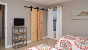 A bedroom with a bed, a TV on a stand, a ceiling fan, and a view of the sea through a window. - Beachfront Renovated Condo | Walkable to Dining, Shopping, & Family Fun! | Pool (Gulf Shores)
