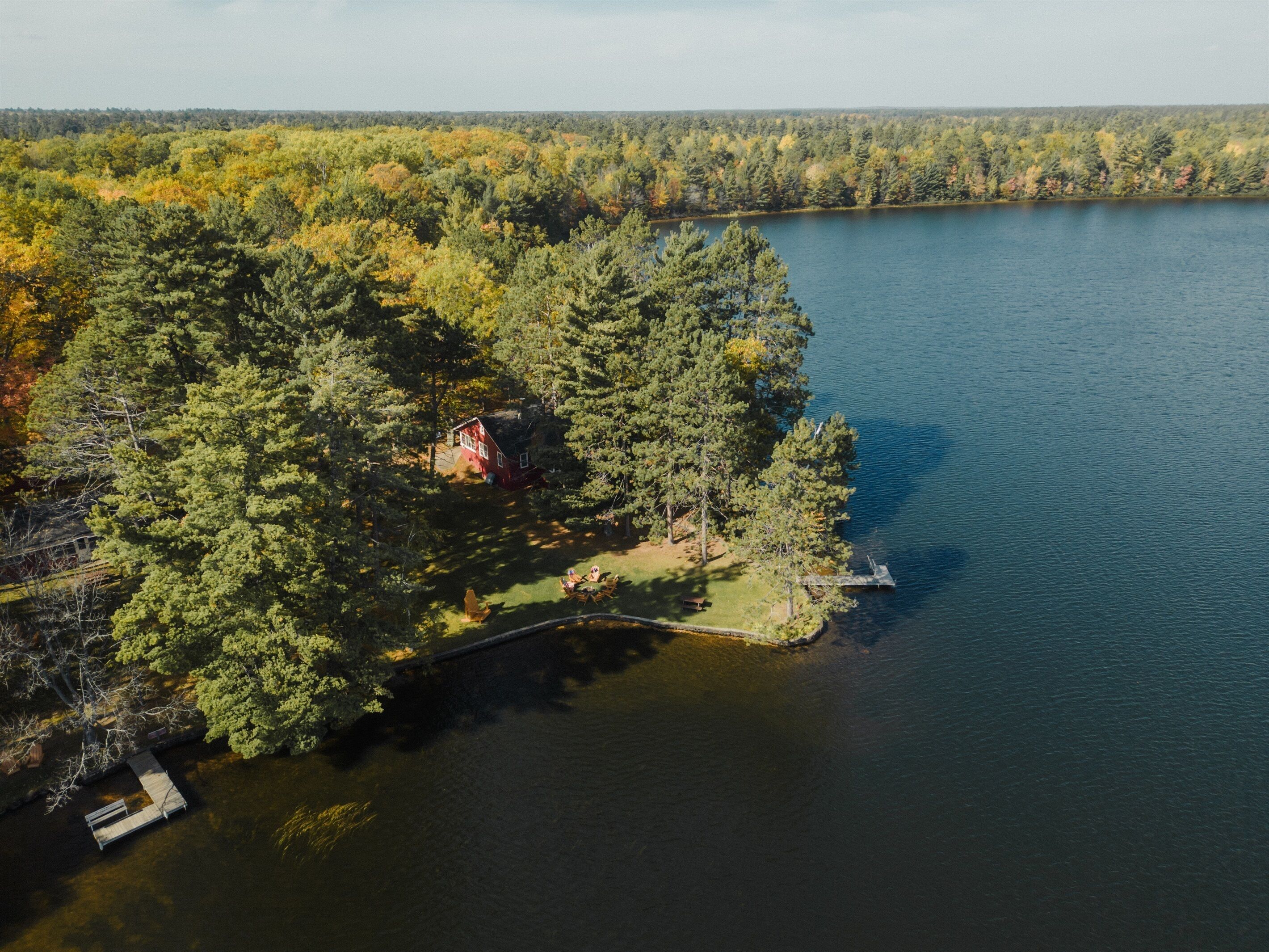 Une vue aérienne d’une propriété au bord du lac, avec un quai, une petite maison entourée d’une forêt dense et une vue dégagée sur le lac.