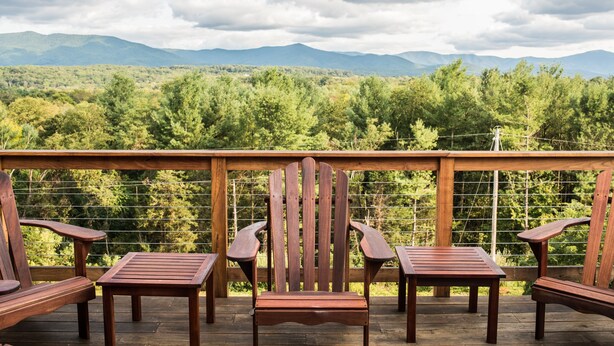 Eine Holzterrasse mit Adirondack-Stühlen und einem Tisch, mit Blick auf einen bewaldeten Gebirgszug.