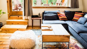 Ein gemütliches Wohnzimmer mit einer Couch, einem Couchtisch und einem flauschigen weißen Hocker. Eine Holztreppe führt zu einer Außenterrasse mit Stühlen.