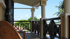 Uma área externa com assento, uma cadeira de madeira e uma mesa preta, um alpendre coberto com vista para áreas verdes e uma palmeira, e entrada de um edifício com uma porta preta.