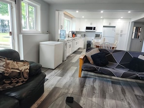 A modern kitchen with white cabinets, a built-in microwave, and a refrigerator. There is a dining area with a table and chairs, and a living room with a sofa and a coffee table. - Unique Air-Conditioned Balsam Lake Waterfront,5 Bedrooms, 3 Baths, Dog friendly. (Kirkfield)