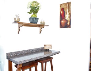 A kitchen with a granite countertop, wooden stools, a hanging plant shelf, and a decorative sign.