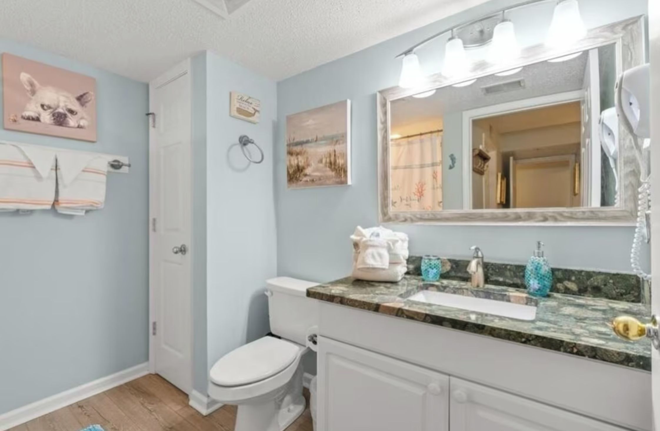 A bathroom with a toilet, sink, and mirror. There are towels hanging on the wall and a picture of a dog.