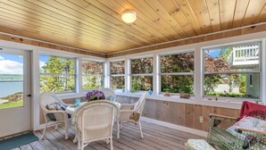 A sunroom with wooden ceiling, large windows, wicker furniture, and a view of a lake.
