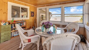 A cozy room with a wooden ceiling, a round glass table with chairs, a bookshelf, and a window with a view of the beach.
