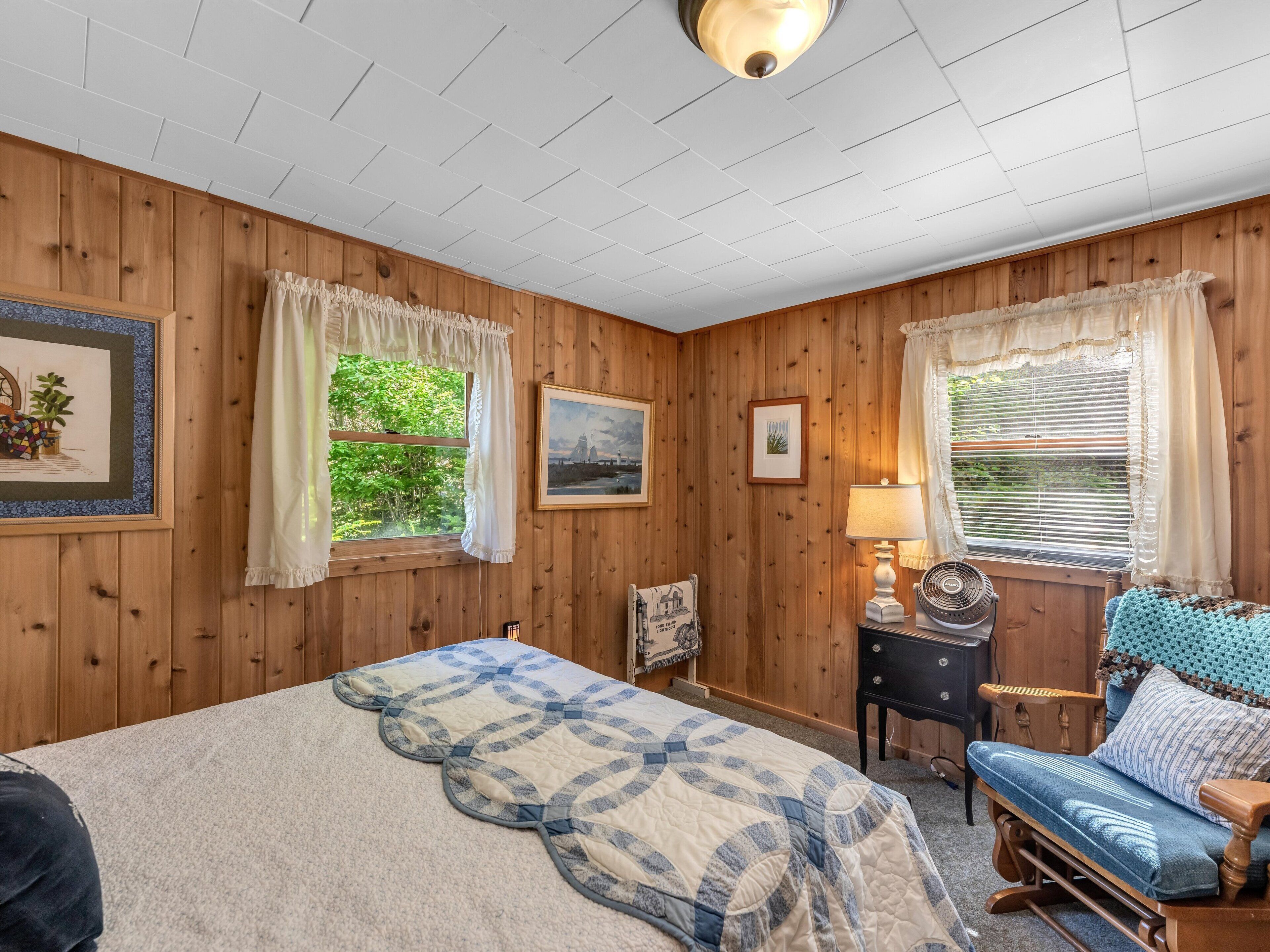 A cozy room with wooden walls, a bed with a quilt, a chair, a lamp, and two windows with curtains.