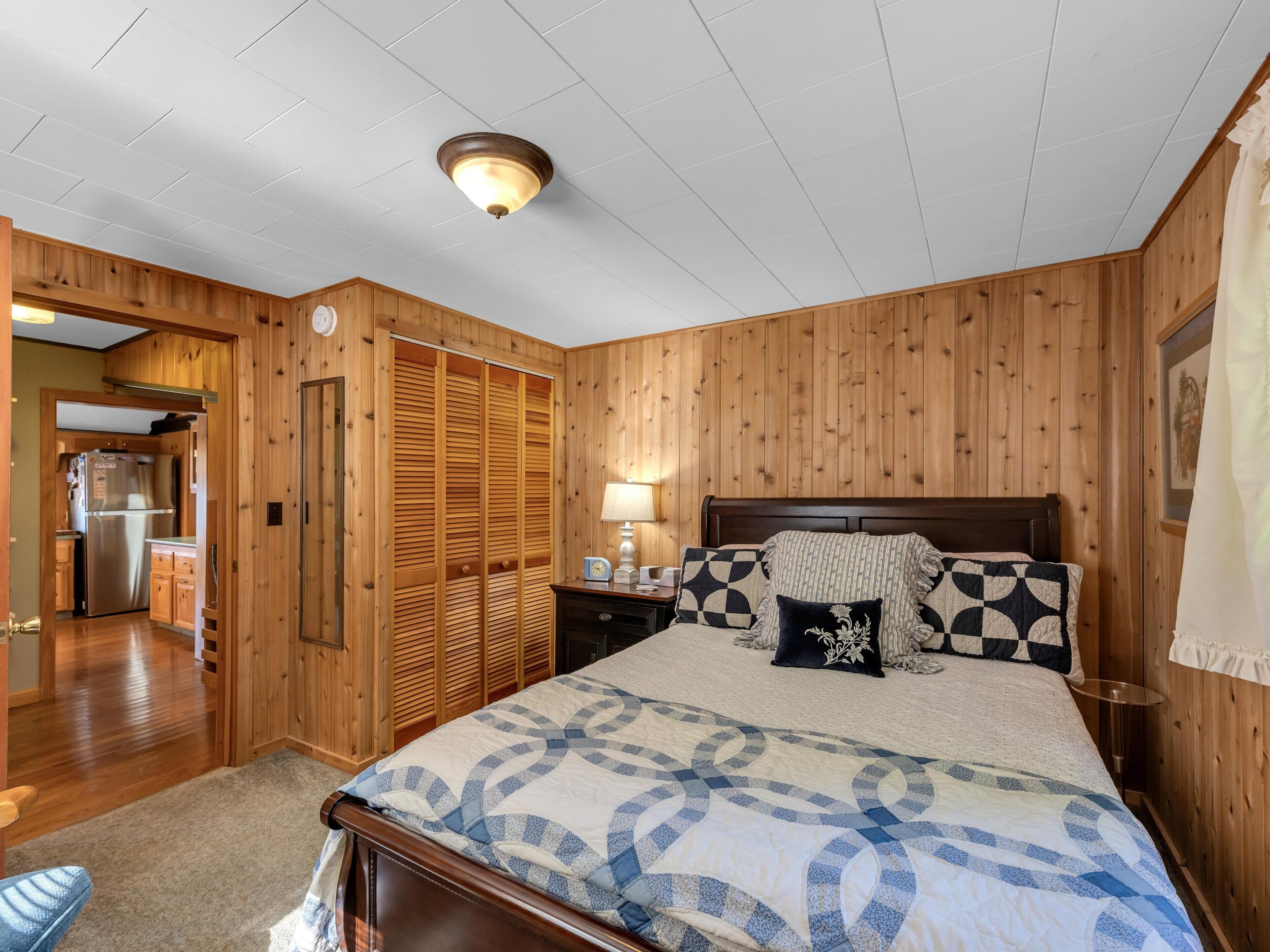 Ein Schlafzimmer mit einer holzverkleideten Wand, einem Bett mit gemustertem Überwurf, einem Nachttisch mit Lampe und einem Holzschrank.